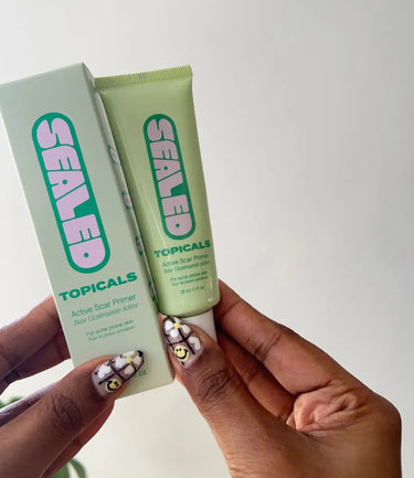 Scar removal cream held by a person with floral nail art against a neutral background.